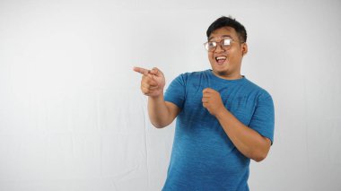 Gözlüklü, mavi tişörtlü, kot pantolonlu genç adam parmaklarını beyaz stüdyo arka planına yaslamış şekilde poz veriyor ve el kol hareketi yapıyor..