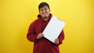 Genç mutlu Asyalı adam beyaz tişört giyiyor ve beyaz bir işaret gösteriyor ve sarı arka planda izole edilmiş kamerada gülümsüyor.