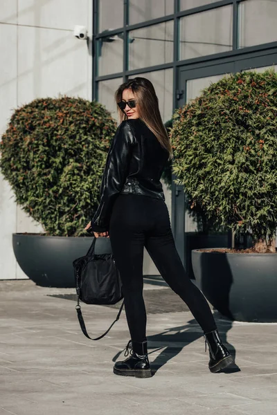 时尚的年轻女性 身穿紧身长袖紧身衣 身穿一身黑色皮夹克 脚跟平整 站在城市街道上 — 图库照片