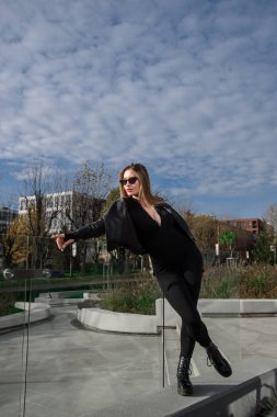 Uzun saçlı, uzun kollu sıkı bir vücut giyen şık bir genç kadın. Siyah deri ceket ve düz platform botlar giymiş bir tulum.