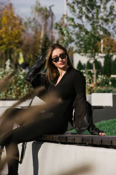 城市妇女坐在公园的长椅上 呼吸着新鲜的空气 头戴长发 身穿固体长袖紧身衣 身穿黑色皮夹克的女人 — 图库照片