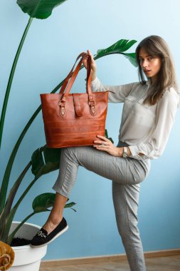 Bir kadının elindeki turuncu deri çantanın yakın plan fotoğrafı. İç mekan fotoğrafı. Gri ekose pantolonlu, beyaz bluzlu, el çantasıyla poz veren güzel, ince bir kız.