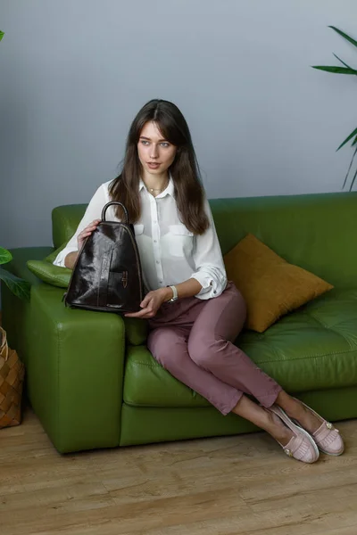 穿着勃艮第裤子的美丽纤细的女孩 穿着一件白色的衬衫 坐在一个绿色沙发上 背着一个小的闪亮的棕色皮背包 室内照片 — 图库照片