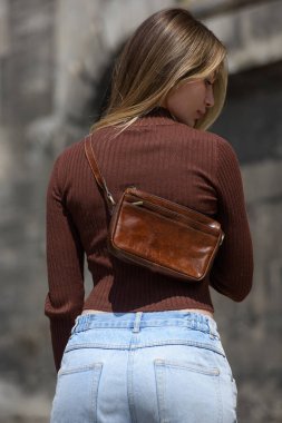 Küçük kahverengi deri çantanın yakın plan fotoğrafı. Açık hava fotoğrafı. Kahverengi gömlekli ve mavi kotlu kız.