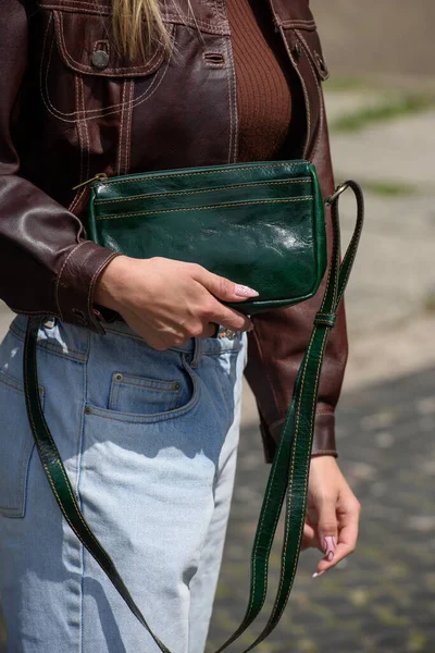 close-up photo of small green leather bag in a woman hands. outdoors photo. Girl in a brown shirt and blue jeans