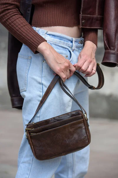 close-up photo of small brown leather bag in a woman hands. outdoors photo. Girl in a brown shirt and blue jeans