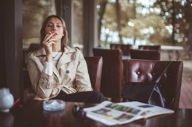 Beautiful woman sitting alone in cafeteria, smoking a cigarette and drinking coffee. Autumn mood