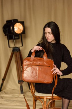 a brunette girl in a knitted black dress poses while standing with a shiny red leather backpack in her hands