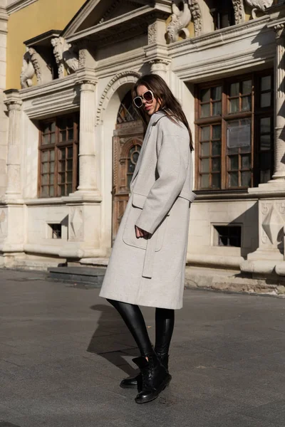 Street style, autumn, spring fashion concept: fashionable woman wearing luxury beige coat, sunglasses, wide leg black leggings, ankle bots. Full length shot on beige background.