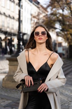 İlkbaharda sokakta poz veren çekici bir esmer, şık bir bej ceket, ustura ve güneş gözlüklü bir bluz giyiyor.