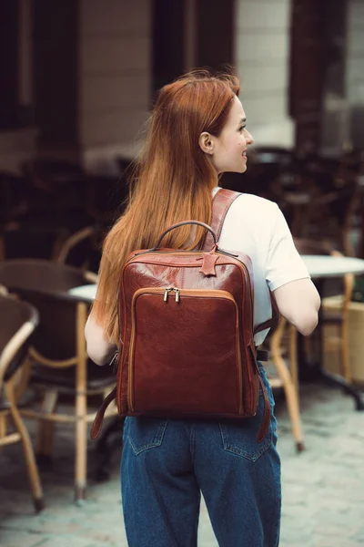 Stock image beautiful red-haired girl with red lips posing with a orange leather backpack. wearing blue jeans and a sweater