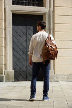 Lüks deri sırt çantasıyla sokakta yürüyen yakışıklı bir iş adamı. Mavi pantolon, spor ayakkabı, beyaz tişört ve bej gömlek giyen bir adam.