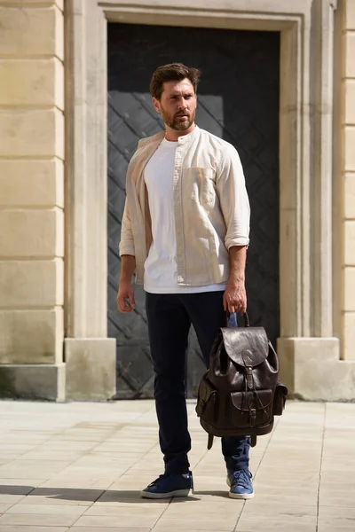 ハンサムなビジネスマンは 高級レザーバックパックで通りを歩いています ブルーパンツ スニーカー ホワイトTシャツ ベージュシャツを着ている男性 — ストック写真