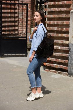 Kot pantolon, gömlek ve beyaz tişört giyen, siyah deri sırt çantalı güzel bir genç kız. Sokak fotoğrafı