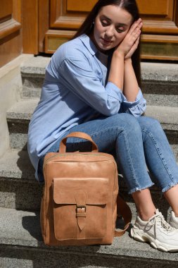 Mavi kot pantolon, gömlek ve beyaz tişört giyen güzel bir genç kız açık kahverengi deri sırt çantasıyla poz veriyor. Sokak fotoğrafı