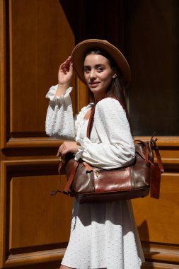 Esmer, uzun saçlı, beyaz elbiseli, bej şapkalı, lüks deri çantalı. Ahşap arka plan. Tatil, turizm ve seyahat