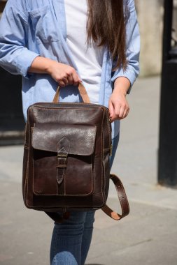 Kot pantolon, gömlek ve beyaz tişört giyen güzel bir genç kız kahverengi deri sırt çantasıyla poz veriyor. Sokak fotoğrafı