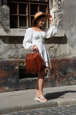 Esmer, uzun saçlı, beyaz elbiseli, bej şapkalı, lüks deri çantalı. Eski şehir sokağı. Tatil, turizm ve seyahat