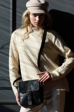 Siyah çantalı, gri arka planda poz veren kıvırcık sarı saçlı güzel bir kadının stüdyo portresi. Şık bir şapka, süveter ve klasik pantolon giyiyor.