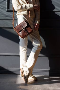 Gri duvarın yanında küçük kahverengi bir çantayla poz veren güzel bir kadın. Şık beyaz kazak ve klasik pantolon giyen bir manken.