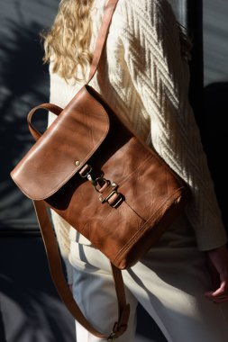 Kıvırcık sarı saçlı, kahverengi sırt çantalı, gri arka planda poz veren güzel bir kadının stüdyo portresi. Şık bir şapka, süveter ve klasik pantolon giyiyor.