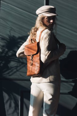 Kıvırcık sarı saçlı, kahverengi sırt çantalı, gri arka planda poz veren güzel bir kadının stüdyo portresi. Şık bir şapka, süveter ve klasik pantolon giyiyor.