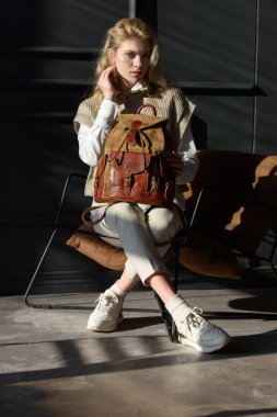 Deri bir sırt çantası tutan bir kadın. Şık örgü yelek, beyaz gömlek ve klasik pantolon giyen bir manken..