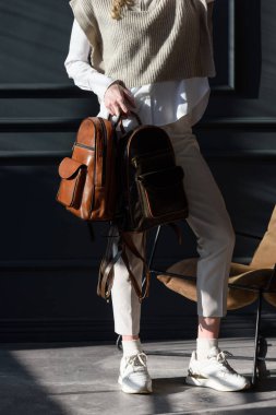 Bir kadın iki kahverengi deri sırt çantası tutar. Şık örgü yelek, beyaz gömlek ve klasik pantolon giyen bir manken..