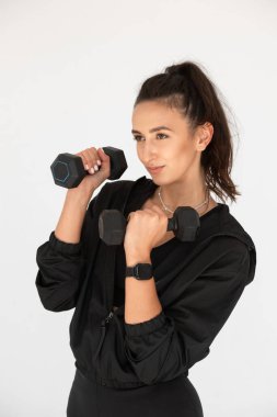 Güç eğitimi için dambıllarla poz veren genç bir kız. Siyah tayt ve gömlek giyen bir sporcu..