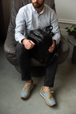 Bir adam yanında sırt çantası olan bir sandalyede rahat bir şekilde oturuyor ve etrafında rahat ve rahat bir atmosfer yaratıyor.