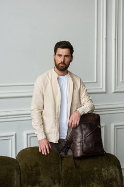 Modern bir odada kahverengi bir çantayla şık bir adam duruyor, kentsel tarzı ve profesyonelliği somutlaştırıyor. Bej gömlek ve kahverengi pantolon