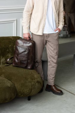Modern bir odada kahverengi bir çantayla şık bir adam duruyor, kentsel tarzı ve profesyonelliği somutlaştırıyor. Bej gömlek ve kahverengi pantolon
