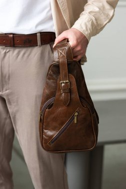 Modern bir odada kahverengi kalça çantasıyla şık bir adam duruyor, şehirli tarzı ve profesyonelliği somutlaştırıyor. Bej gömlek ve kahverengi pantolon