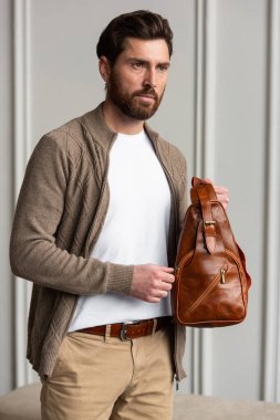 Zarif bir adam sarı kahverengi bir çantayla modern bir odada duruyor, kentsel tarzı ve profesyonelliği somutlaştırıyor. Bej gömlek ve kahverengi pantolon