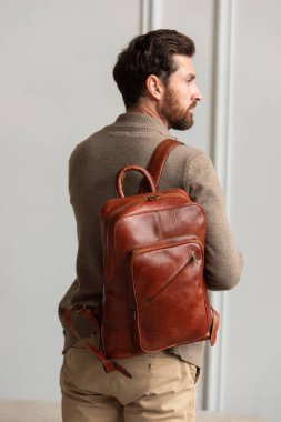 Zarif bir adam sarı kahverengi bir sırt çantasıyla çağdaş bir odada duruyor, kentsel tarzı ve profesyonelliği somutlaştırıyor..