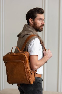 Zarif bir adam sarı kahverengi bir sırt çantasıyla çağdaş bir odada duruyor, kentsel tarzı ve profesyonelliği somutlaştırıyor..