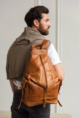 Zarif bir adam sarı kahverengi bir sırt çantasıyla çağdaş bir odada duruyor, kentsel tarzı ve profesyonelliği somutlaştırıyor..