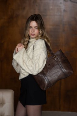 Sıcak kazaklı, siyah kısa etekli, naylon taytlı bir kız kahverengi deri çantayla poz veriyor.
