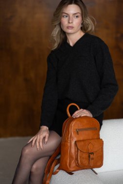 Sıcak kazaklı, siyah kısa etekli ve naylon taytlı bir kız küçük kahverengi bir sırt çantasıyla poz veriyor.