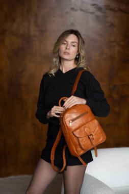 Sıcak kazaklı, siyah kısa etekli ve naylon taytlı bir kız açık kahverengi bir sırt çantasıyla poz veriyor.