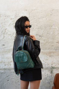 Siyah deri ceket, güneş gözlüğü, ayakkabı ve yeşil bir sırt çantasıyla poz veren kıvırcık kız..