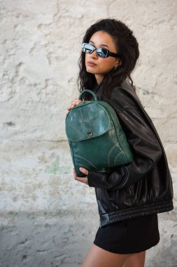 Siyah deri ceket, güneş gözlüğü, ayakkabı ve yeşil bir sırt çantasıyla poz veren kıvırcık kız..