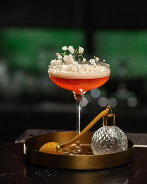 Köpüklü bir üst ile kızarık tonlu kokteyl zarif beyaz bebek nefesli çiçeklerle süslenir, eski tarz parfüm atomizatörünün yanında altın bir tepside sunulur..