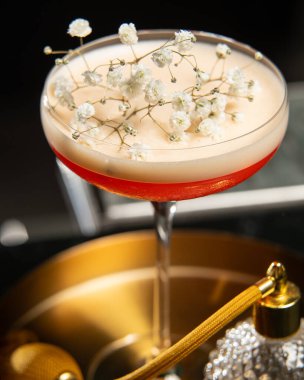 Köpüklü bir üst ile kızarık tonlu kokteyl zarif beyaz bebek nefesli çiçeklerle süslenir, eski tarz parfüm atomizatörünün yanında altın bir tepside sunulur..