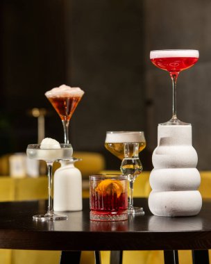 Canlı turuncu kabuklu zengin bir kırmızı kokteyl, loş ışıklı bir barda diğer sofistike içkilerle çevrili, dokulu bir taş bardakta sunuluyor.