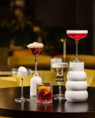 Canlı turuncu kabuklu zengin bir kırmızı kokteyl, loş ışıklı bir barda diğer sofistike içkilerle çevrili, dokulu bir taş bardakta sunuluyor.