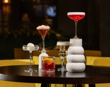 Canlı turuncu kabuklu zengin bir kırmızı kokteyl, loş ışıklı bir barda diğer sofistike içkilerle çevrili, dokulu bir taş bardakta sunuluyor.