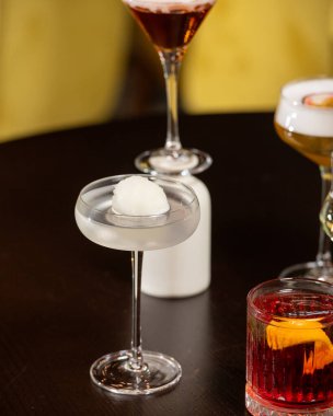 Canlı turuncu kabuklu zengin bir kırmızı kokteyl, loş ışıklı bir barda diğer sofistike içkilerle çevrili, dokulu bir taş bardakta sunuluyor.