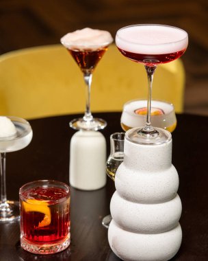 Canlı turuncu kabuklu zengin bir kırmızı kokteyl, loş ışıklı bir barda diğer sofistike içkilerle çevrili, dokulu bir taş bardakta sunuluyor.