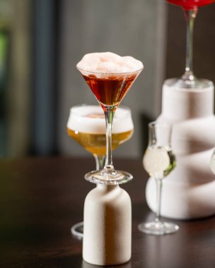 Canlı turuncu kabuklu zengin bir kırmızı kokteyl, loş ışıklı bir barda diğer sofistike içkilerle çevrili, dokulu bir taş bardakta sunuluyor.